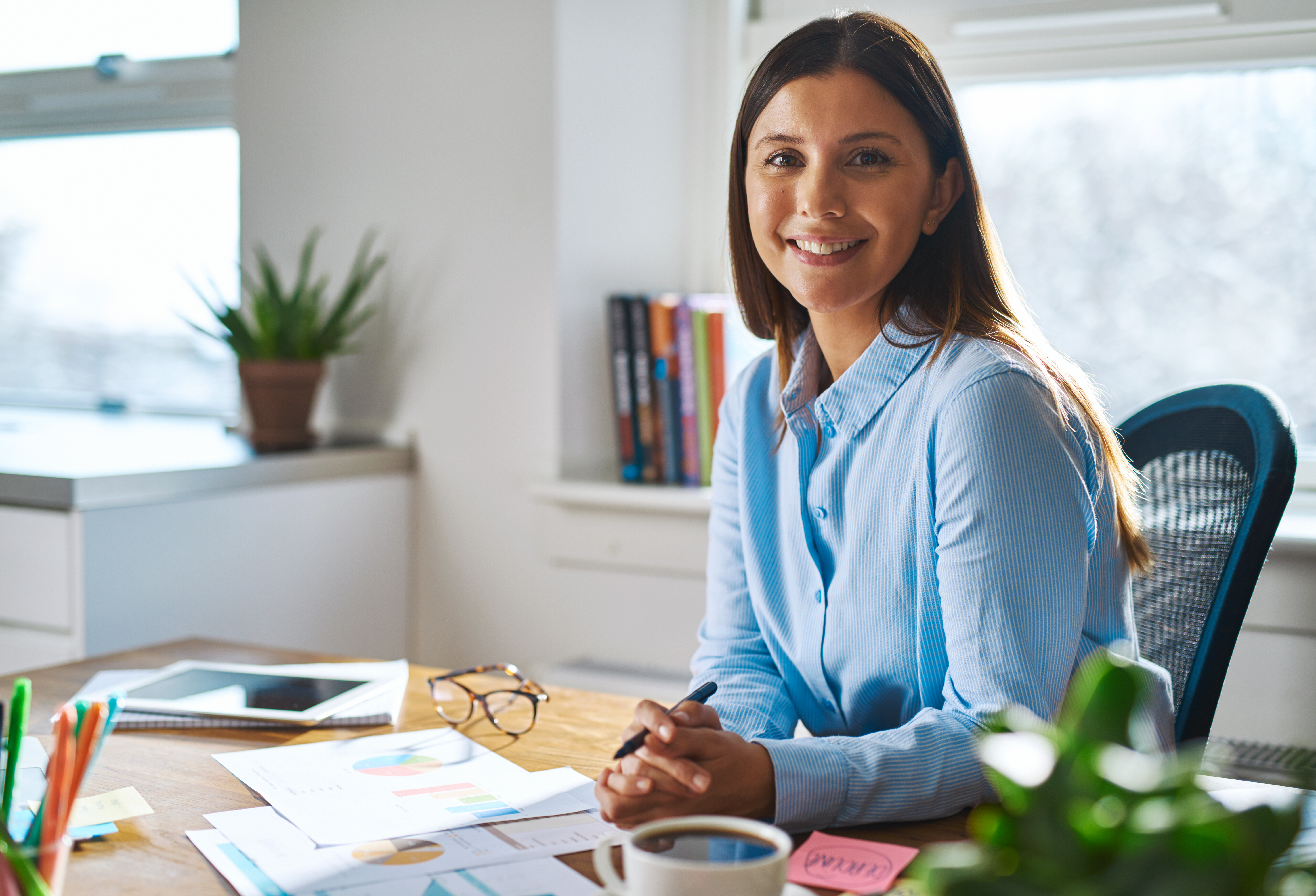 Single confident smiling woman in blue shirt with folded hands at desk in a bright, sunlight filled office